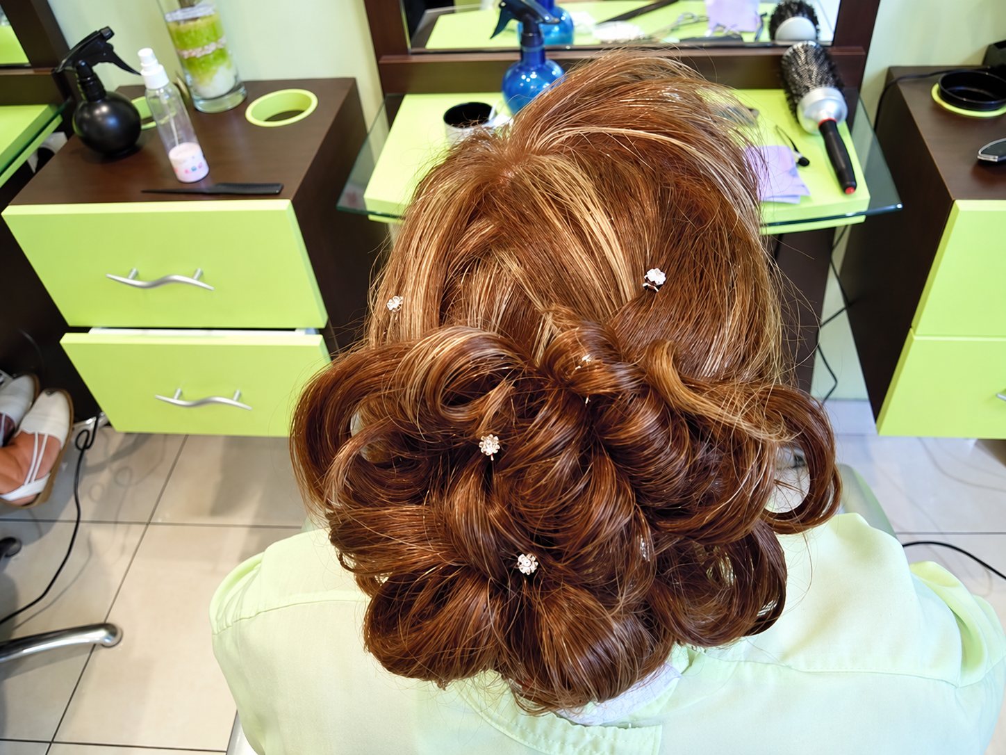 Vue de dessus d'un chignon aux boucles châtain cuivré décoré de petites épingles brillantes, dans un salon de coiffure aux meubles vert anis.