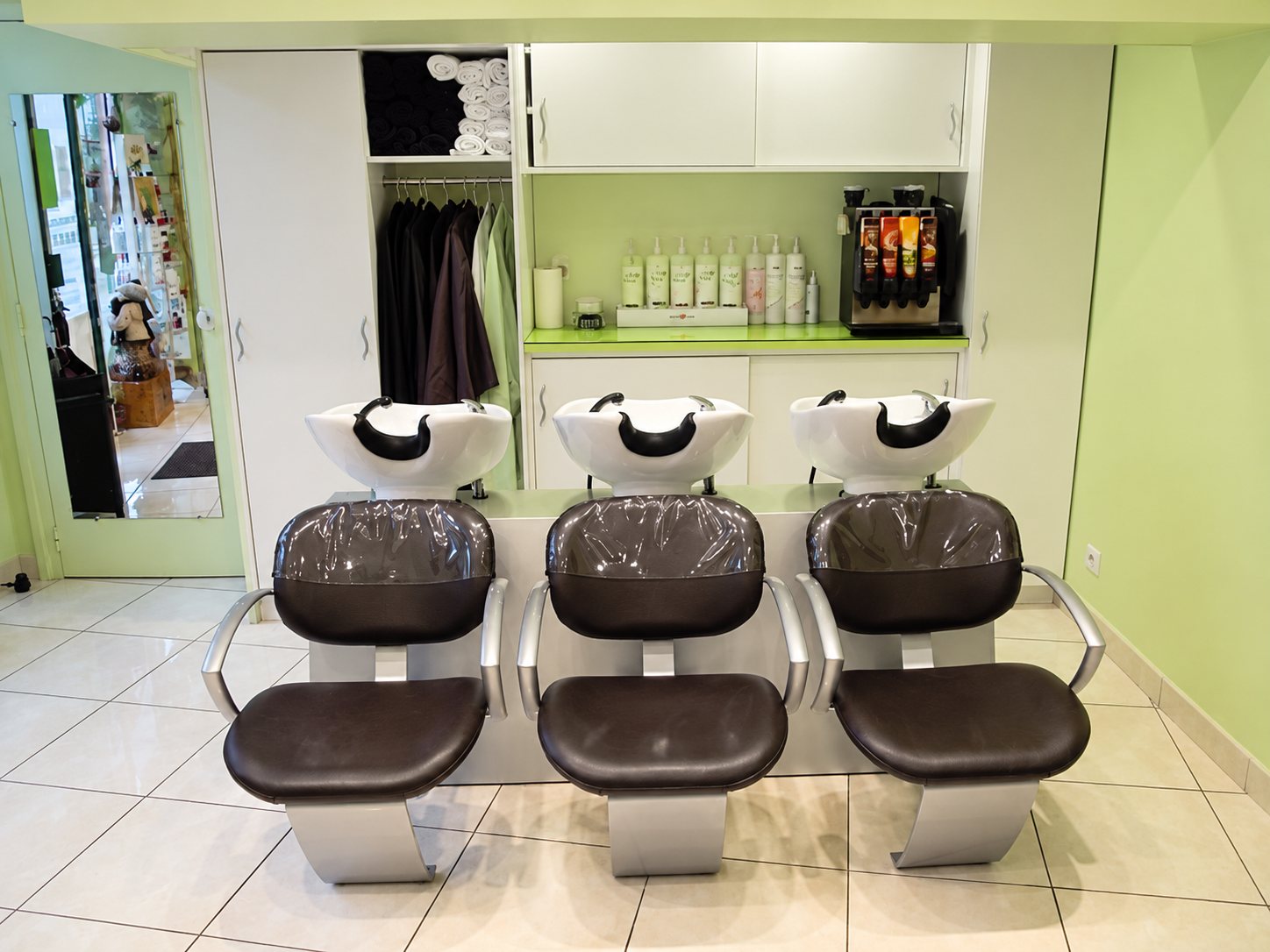 Dans un salon de coiffure aux murs vert clair, trois fauteuils bruns font face à trois bacs de lavage blancs, avec des flacons de produits et des serviettes rangés au-dessus.