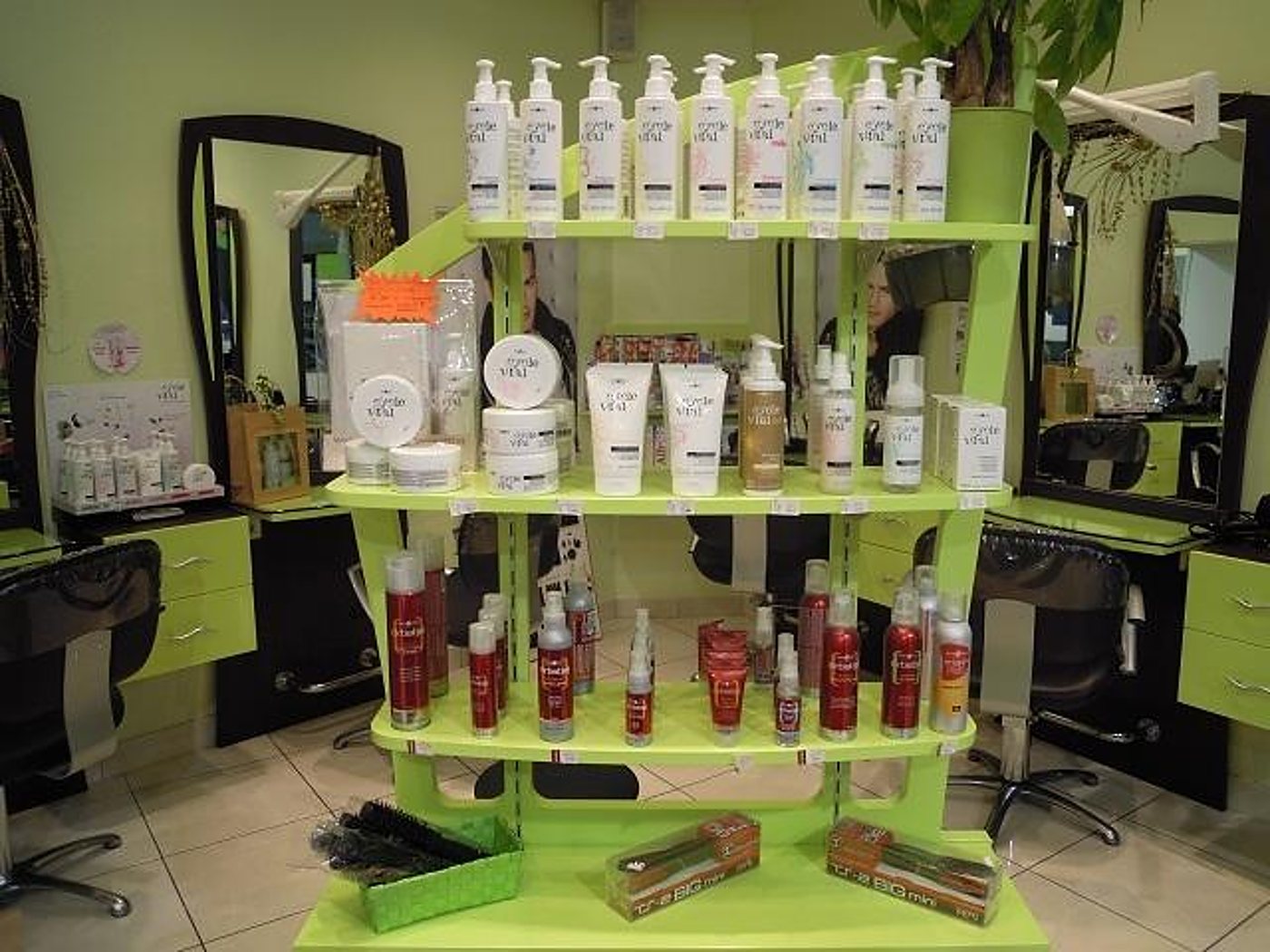 Une étagère verte à trois niveaux présente des flacons de shampooing, des soins et des laques devant les miroirs et les fauteuils d'un salon de coiffure.