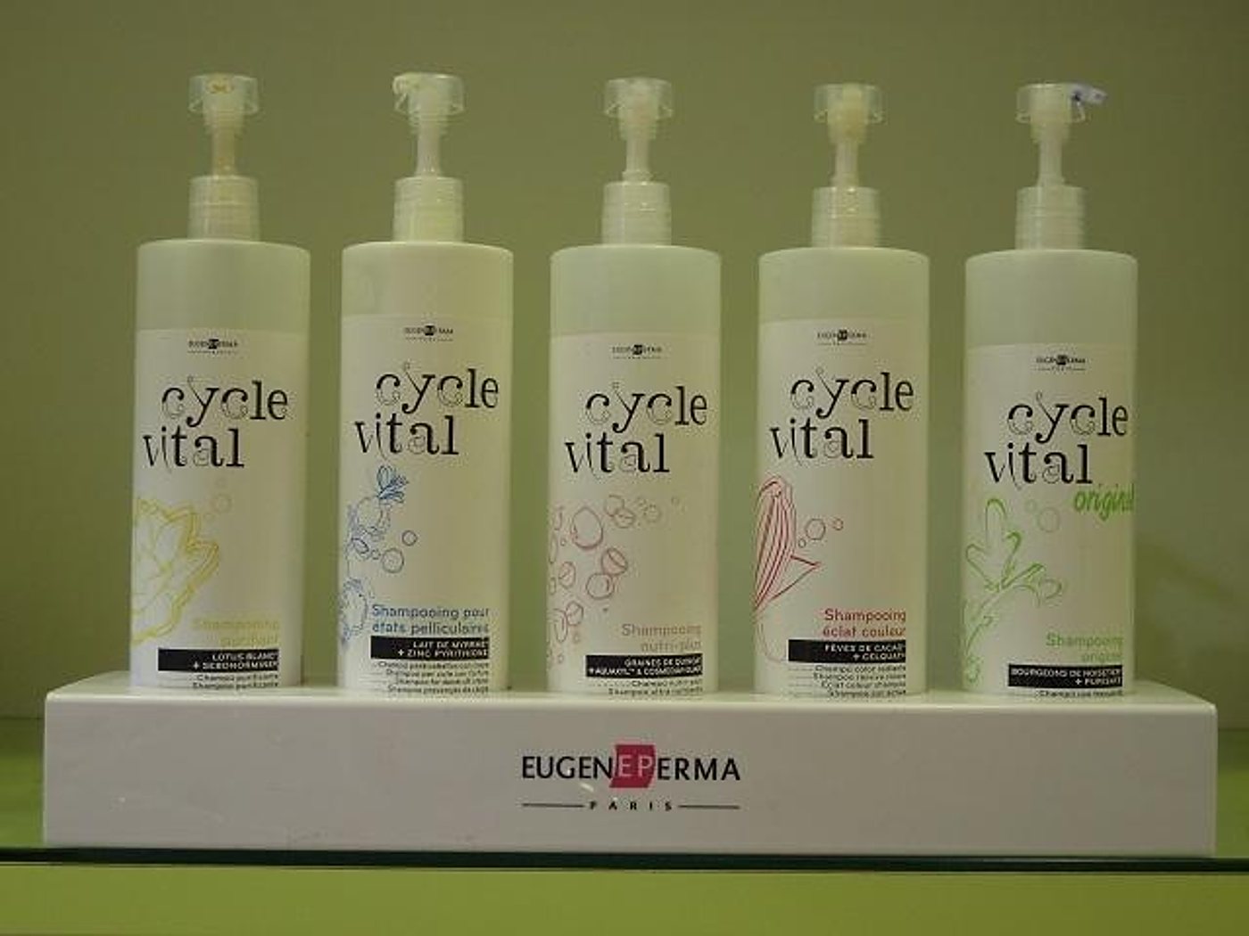 Cinq grands flacons pompe de shampooing Cycle Vital alignés sur un présentoir blanc portant le nom Eugène Perma Paris, devant un mur vert.