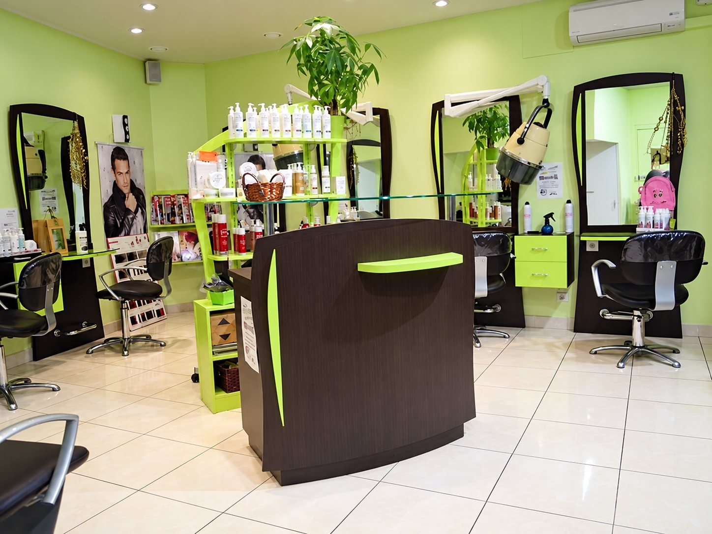 Intérieur d'un salon de coiffure aux murs vert clair, avec comptoir en bois sombre, fauteuils noirs, grands miroirs et étagères de produits capillaires.