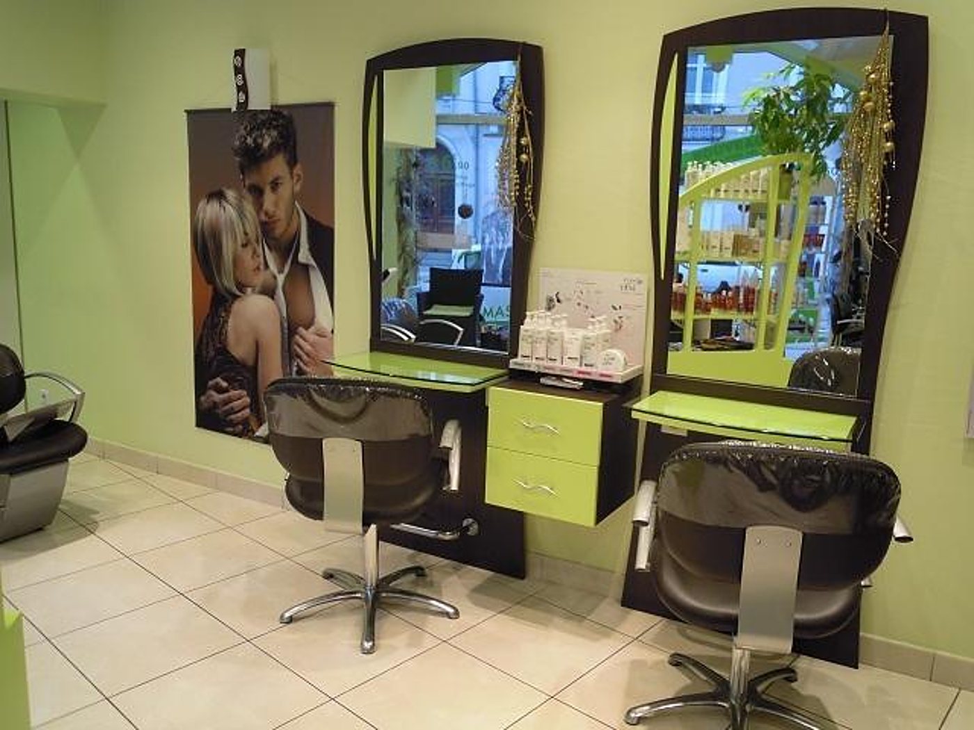 Intérieur d'un salon de coiffure aux murs vert anis, avec deux fauteuils bruns devant des miroirs, des tiroirs verts et une affiche de couple au mur.
