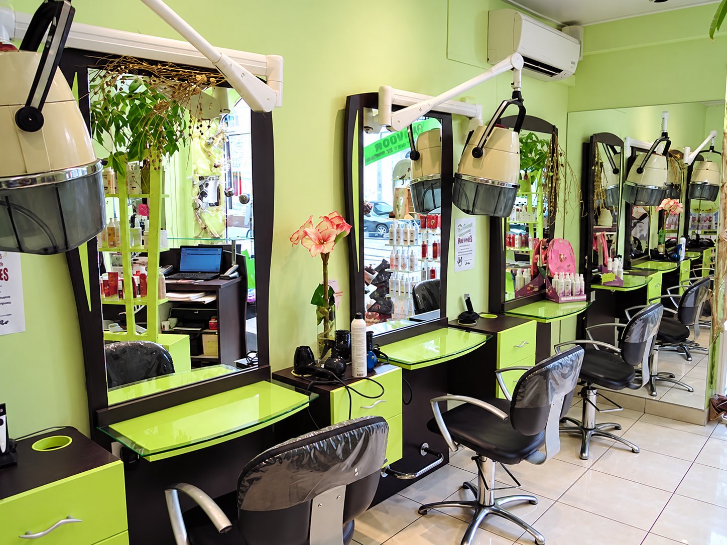 Intérieur d'un salon de coiffure aux murs vert anis, avec des postes de coiffage équipés de miroirs, de casques et de fauteuils noirs.