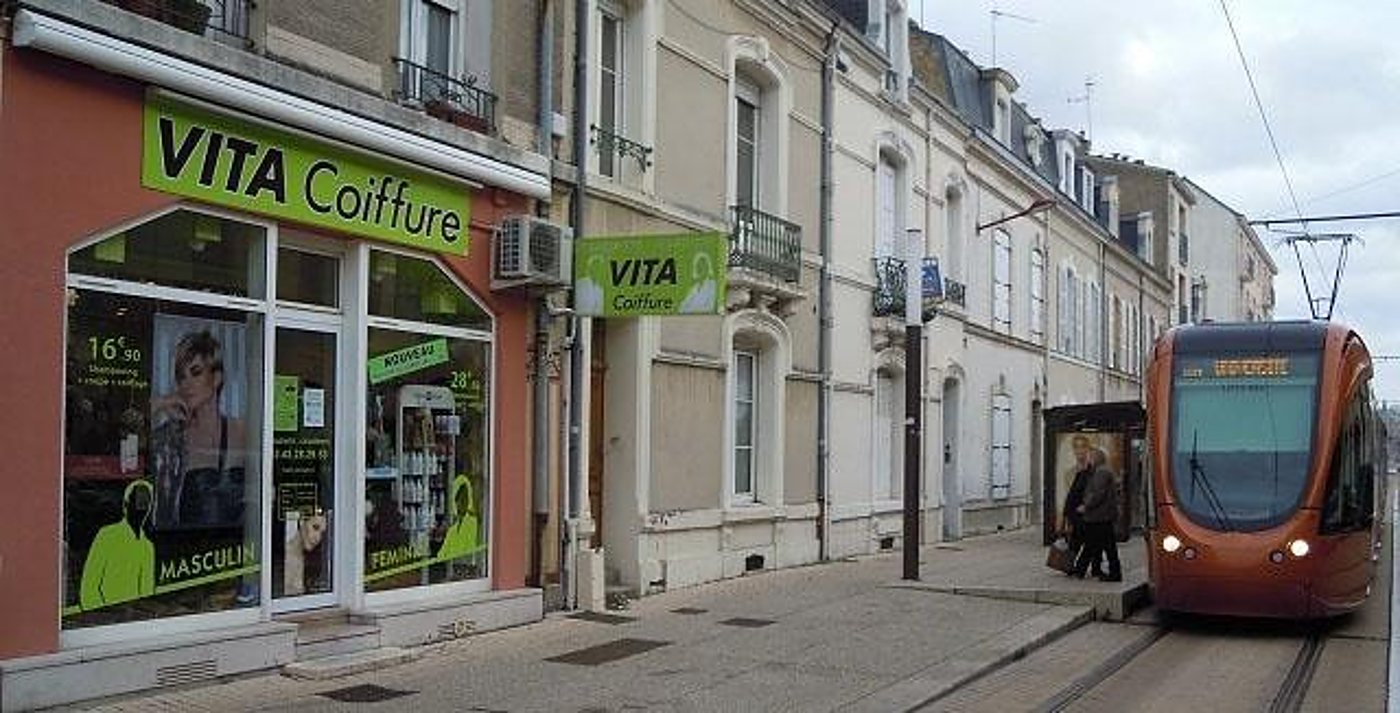 La façade verte et orange du salon de coiffure Vita donne sur une rue où passe un tramway orange.
